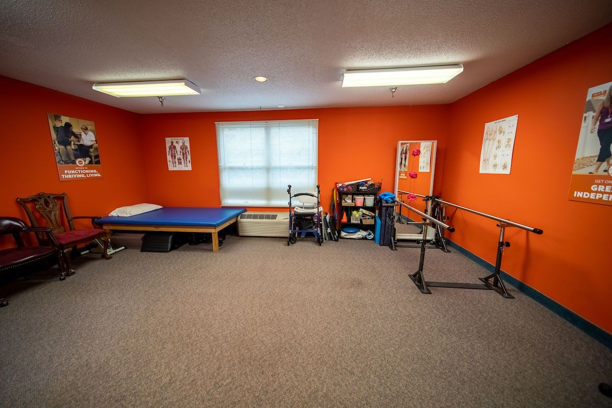 A physical therapy room equipped with parallel bars, a treatment table, and exercise equipment against vibrant orange walls.