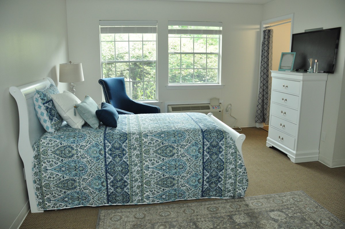 A bright and cozy bedroom featuring a bed with blue patterned bedding, an armchair, and a dresser, all illuminated by natural light from two large windows.