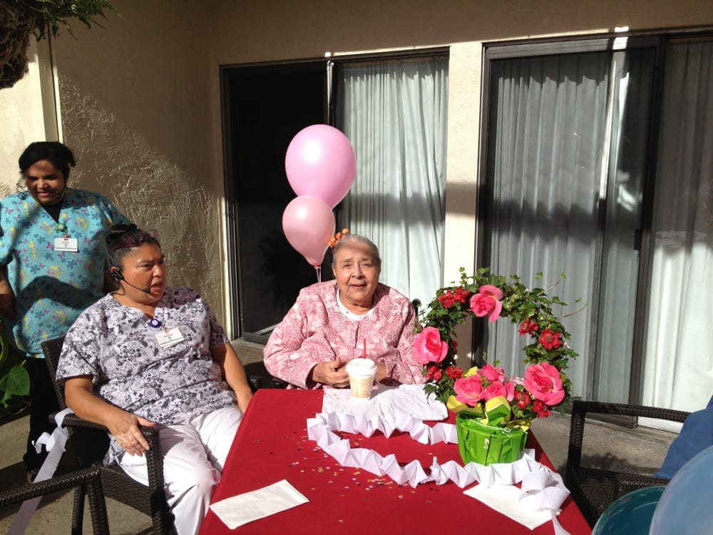 An elderly woman celebrating her birthday outdoors with balloons and flowers, accompanied by another person.