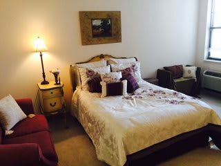 A cozy bedroom interior featuring a neatly made bed with decorative pillows, a bedside table with a lamp, and classic furniture.