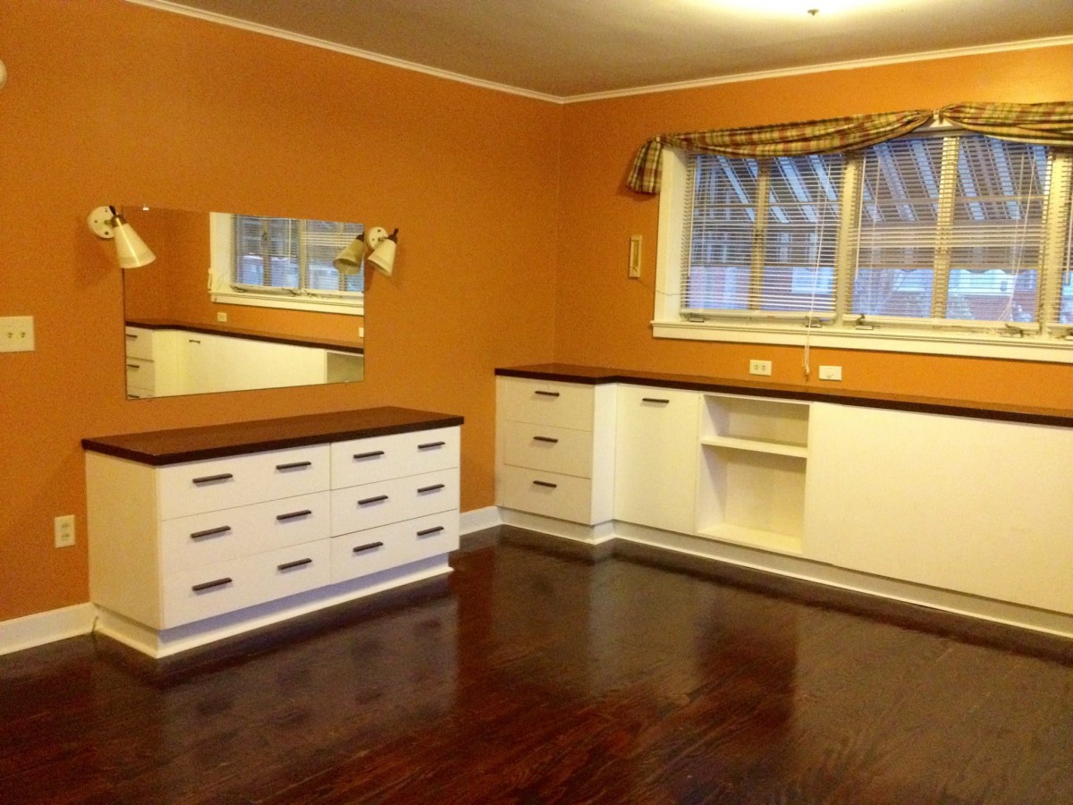 A room with orange walls featuring a white dresser, mirrors, and hardwood flooring.
