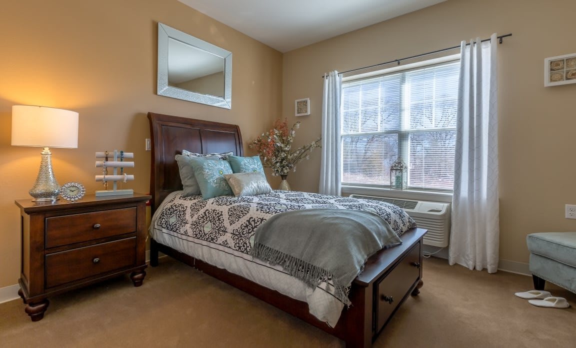 A cozy bedroom with a decorated bed, large mirror, and natural light streaming through the window.