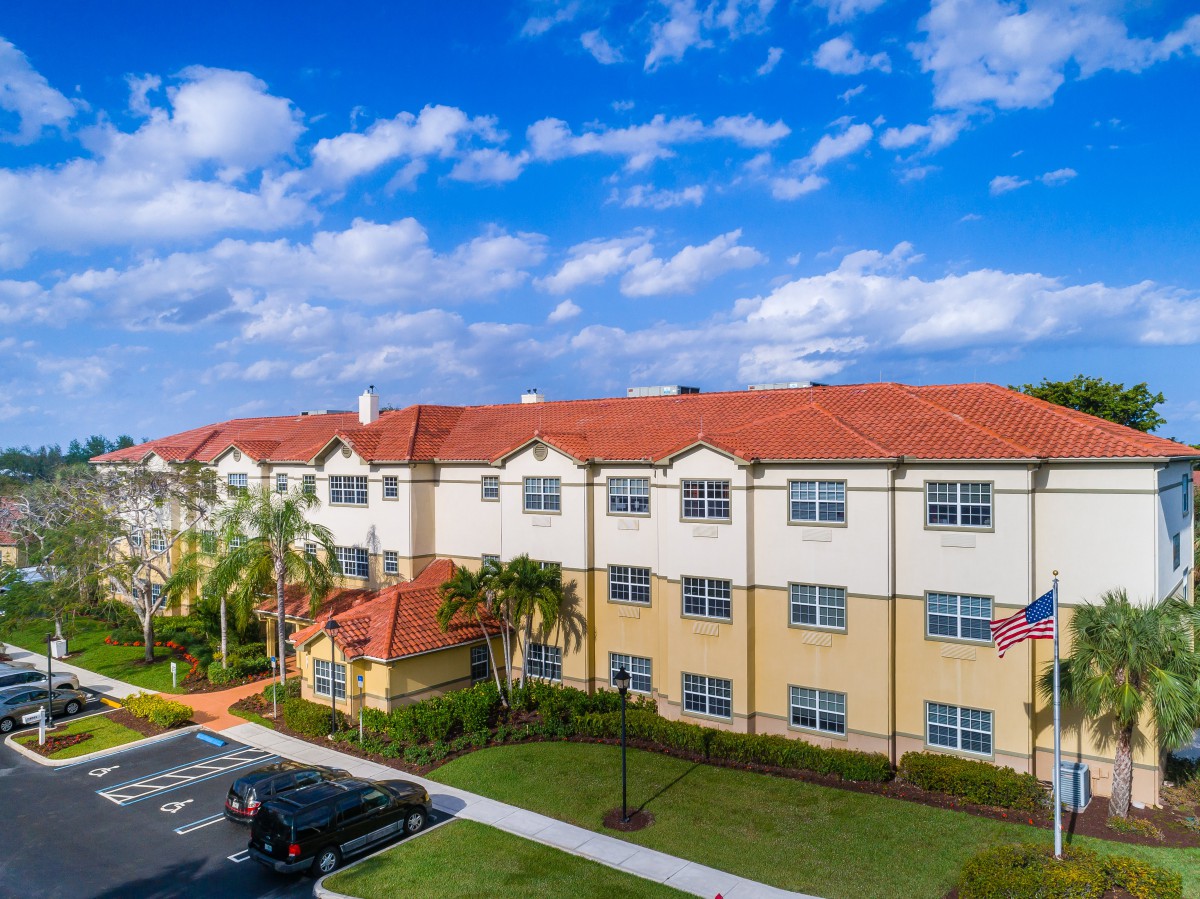 Aerial view of a residential apartment building with a red roof, surrounded by palm trees and a parking area under a partly cloudy sky.
