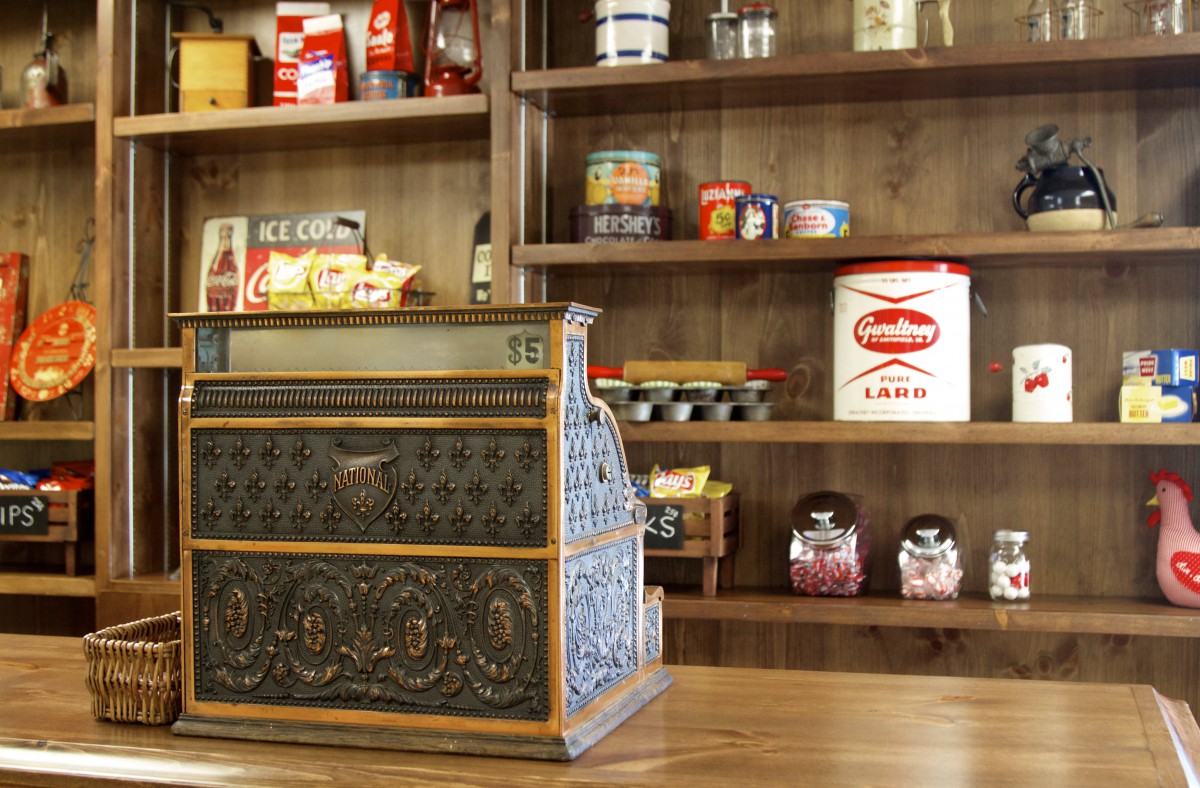 A vintage cash register in an old-fashioned store setting, surrounded by retro items on shelves.