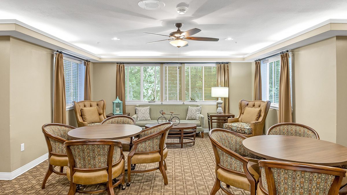 A cozy living room with comfortable seating arrangements, including sofas and chairs around tables, complemented by a ceiling fan and large windows.