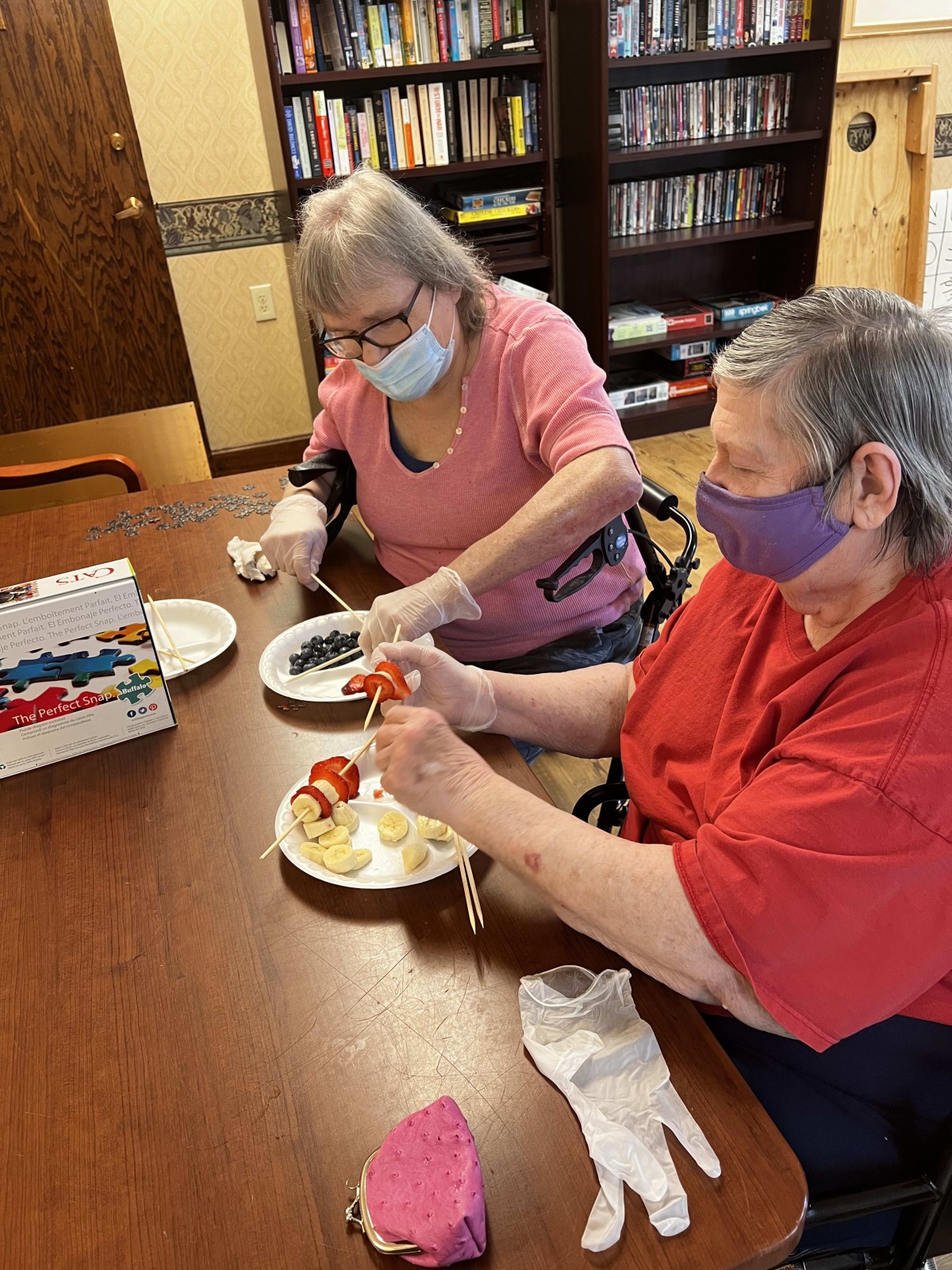 Two elderly women wearing masks are engaged in a crafting activity at a table.