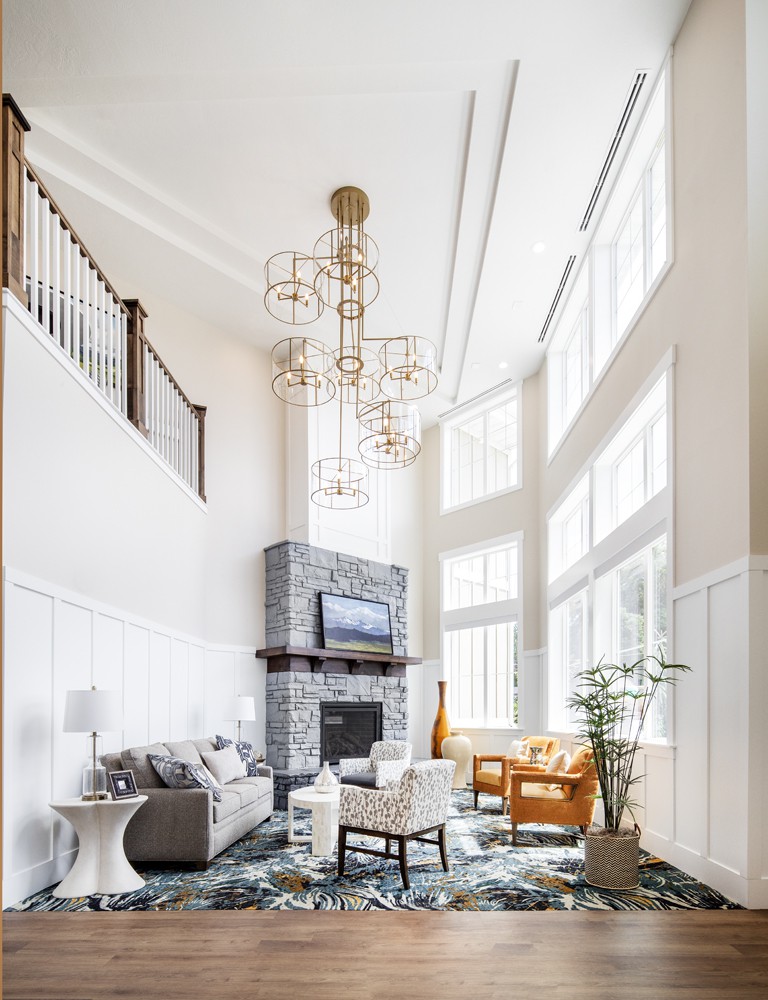 A spacious living room with a high ceiling, large windows, and a modern design featuring a stone fireplace and elegant chandeliers.