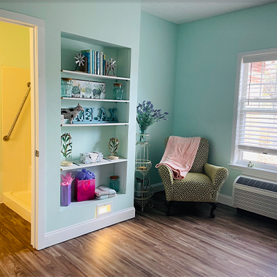 A cozy living room with pastel walls, a shelf filled with decorative items, an armchair by the window, and hardwood flooring.