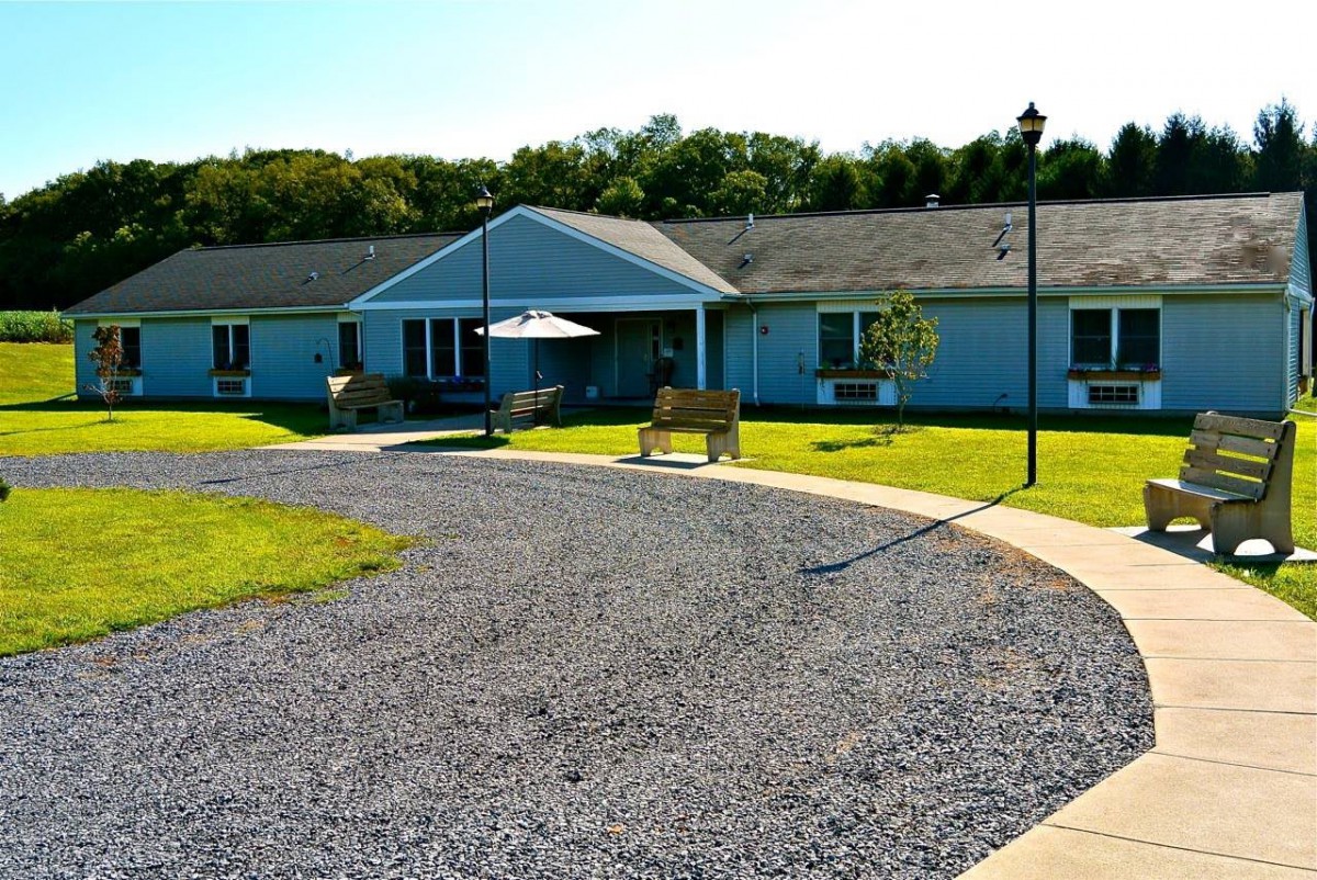 A residential building with a gravel path and a bench in front, surrounded by greenery on a sunny day.