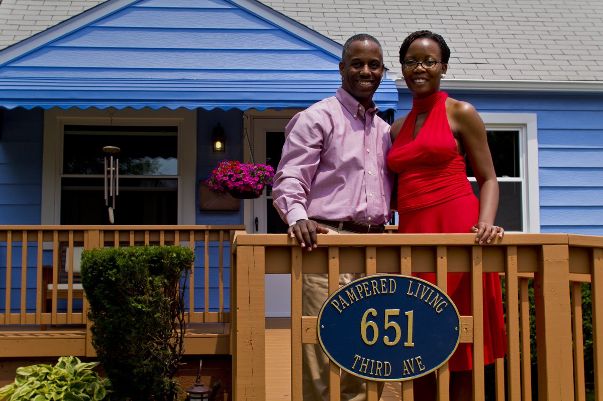 A couple standing on a porch in front of their home, smiling at the camera.