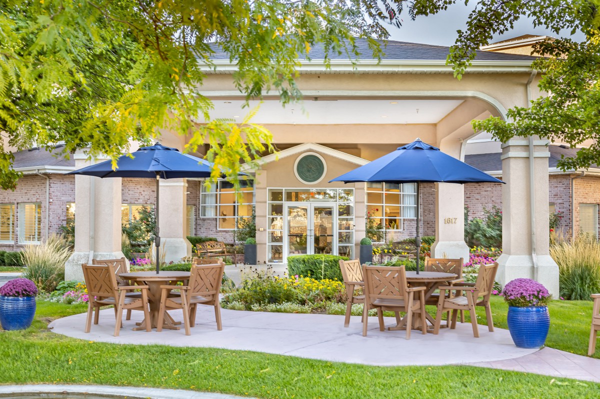 A charming outdoor seating area with tables, chairs, and blue umbrellas set in front of a building surrounded by lush greenery.