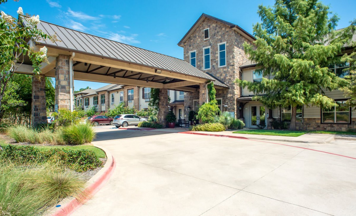 A residential building with a stone exterior, surrounded by greenery and vehicles parked under a carport.