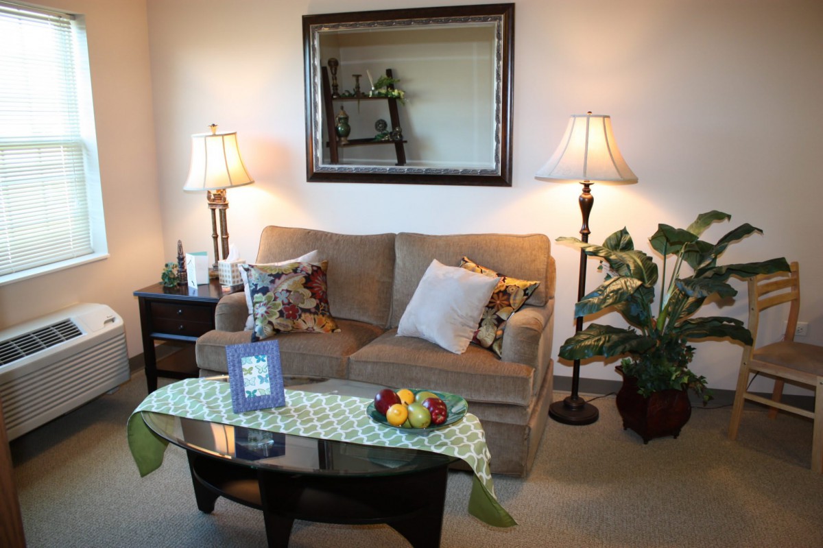 A cozy living room with a sofa, decorative pillows, and a coffee table adorned with fruit and books. Two lamps flank the sofa, adding warmth to the space, while a mirror above reflects the decor.