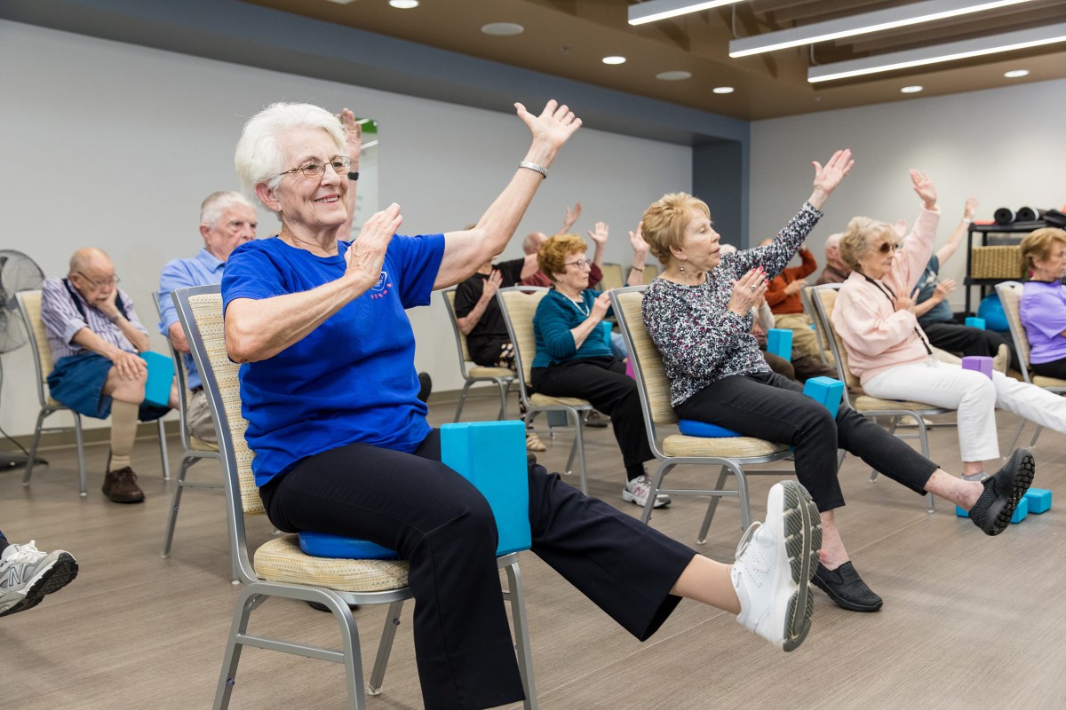 A group of seniors engaging in a seated exercise class indoors, with participants smiling and lifting their arms.