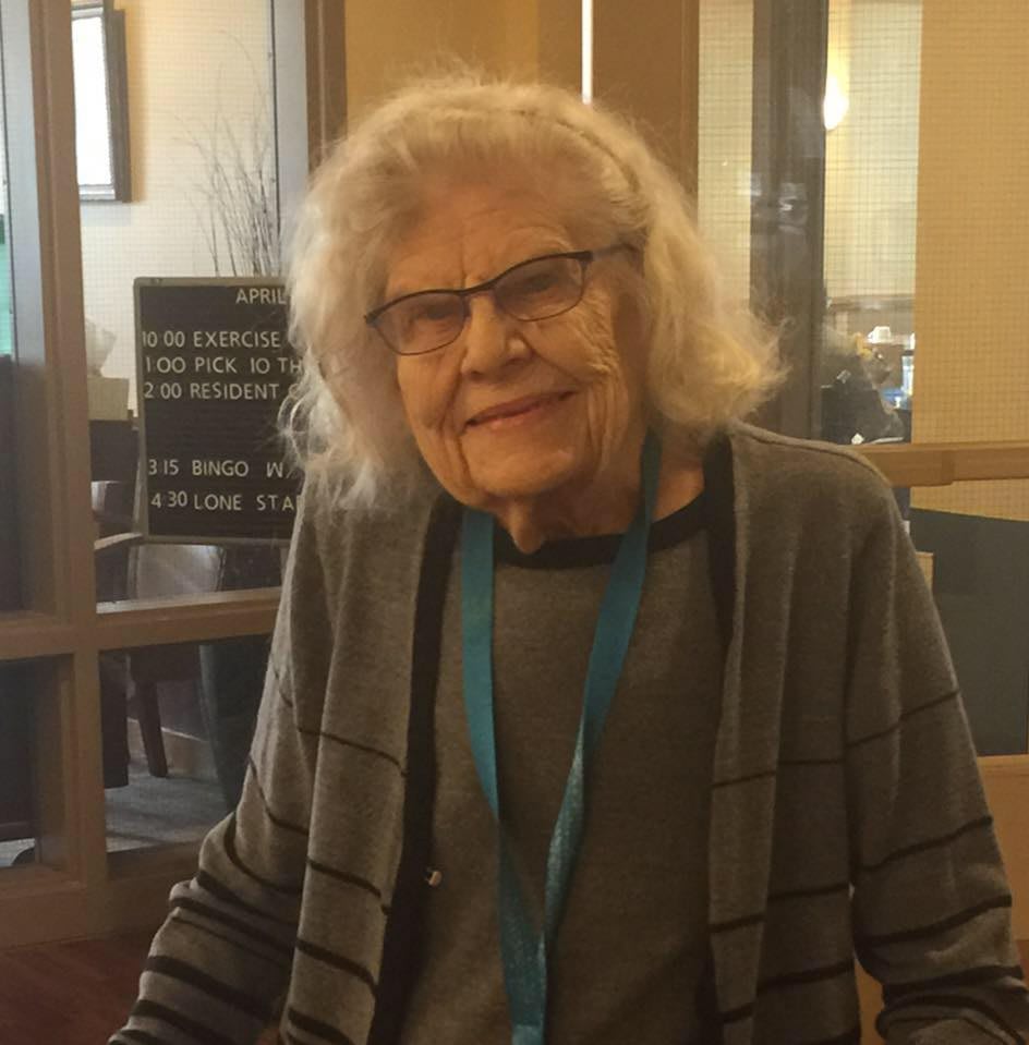 An elderly woman wearing glasses and a lanyard, smiling indoors.
