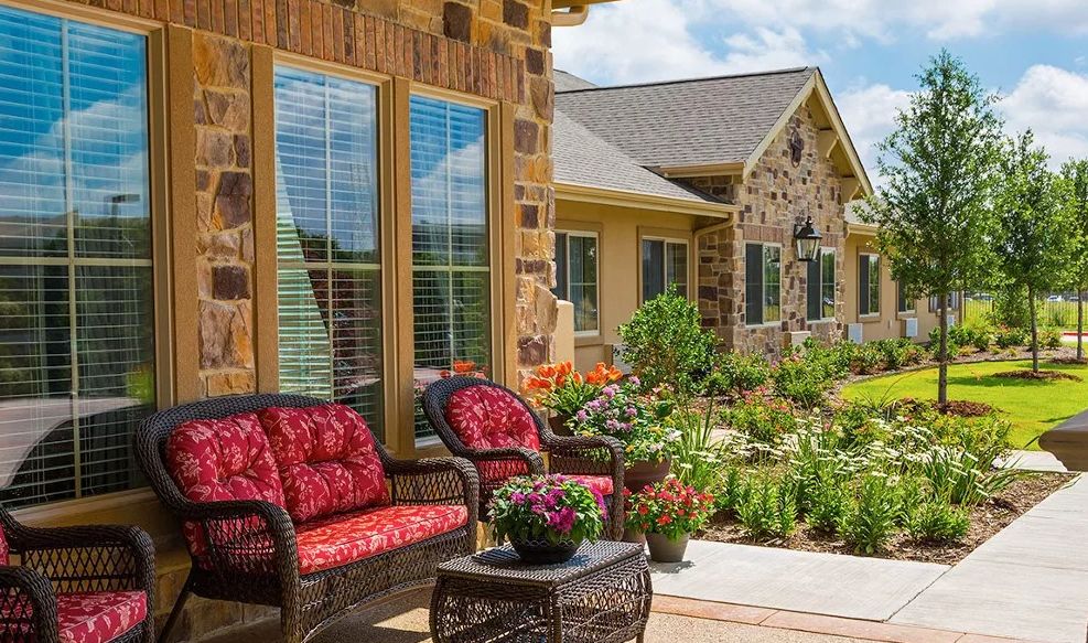Cozy patio area with wicker furniture and colorful cushions in front of a stone house, surrounded by a lush garden.