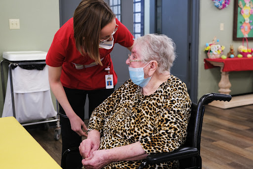 A caregiver attentively interacts with an elderly resident in a nursing home, both wearing masks.