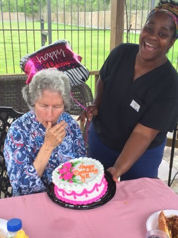 An elderly woman celebrates her birthday with a cake, assisted by a smiling person outdoors.