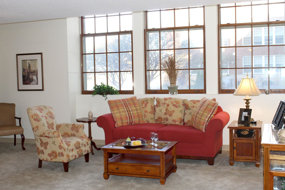 A cozy living room with a red sofa, floral armchair, and wooden coffee table in front of large windows.