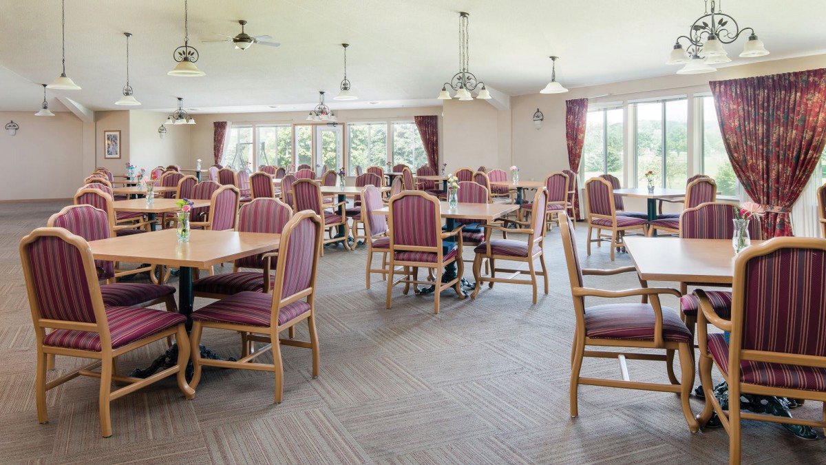 An empty dining room with multiple tables and chairs, featuring large windows and curtains.