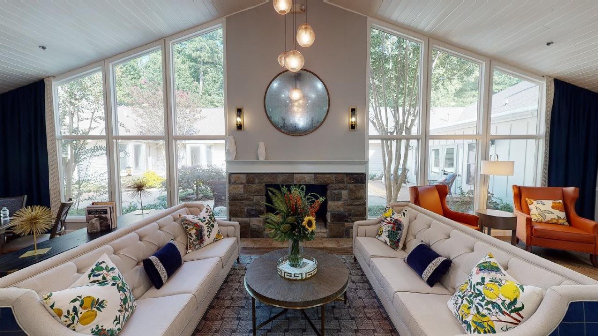 A modern living room with two sofas facing a central coffee table, decorated with floral arrangements. Large windows and a fireplace enhance the bright and cozy atmosphere.