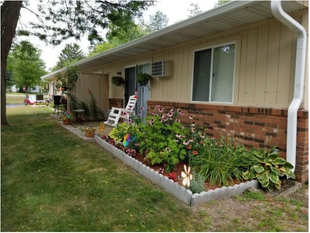 A garden with colorful flowers in front of a brick house, featuring a white rocking chair on the porch.
