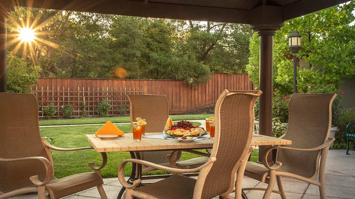 A cozy outdoor patio setup with a table and four chairs, featuring food and drinks, in a lush backyard garden.