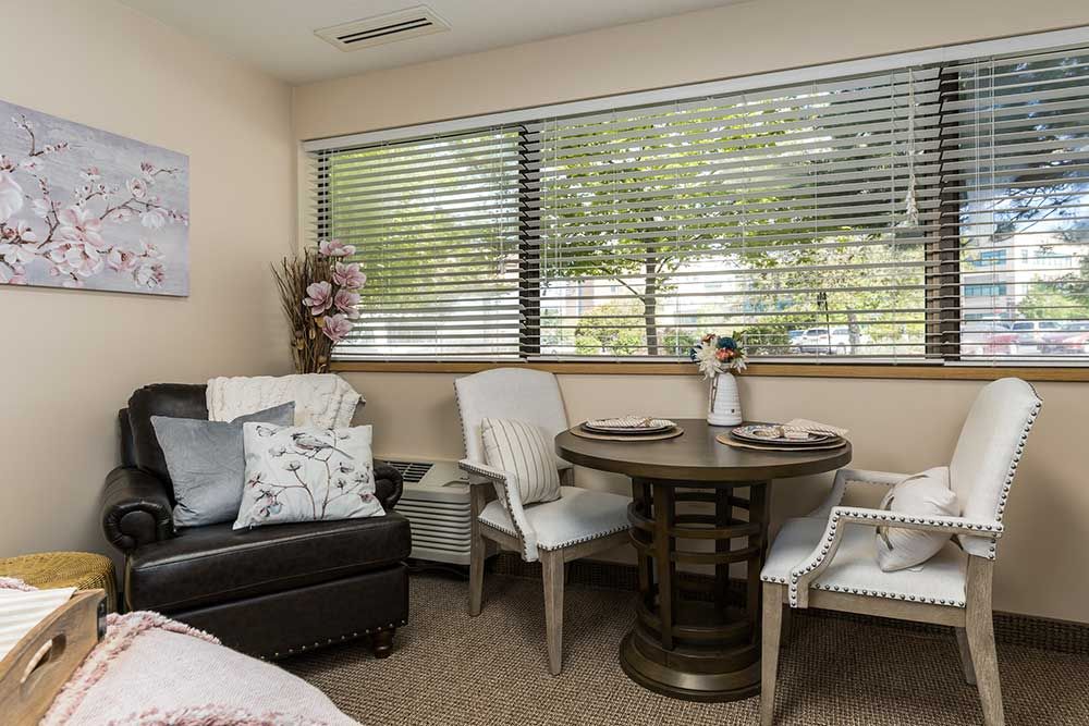 A cozy living room corner featuring a round table with chairs, a comfortable sofa, and large windows with blinds.