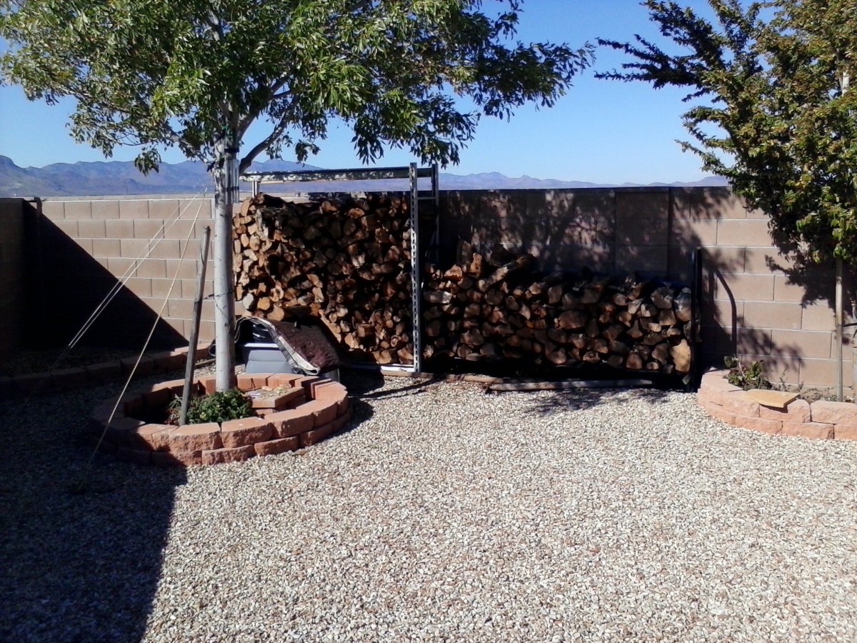 A backyard with a wood pile, trees, and solar panel on gravel ground.