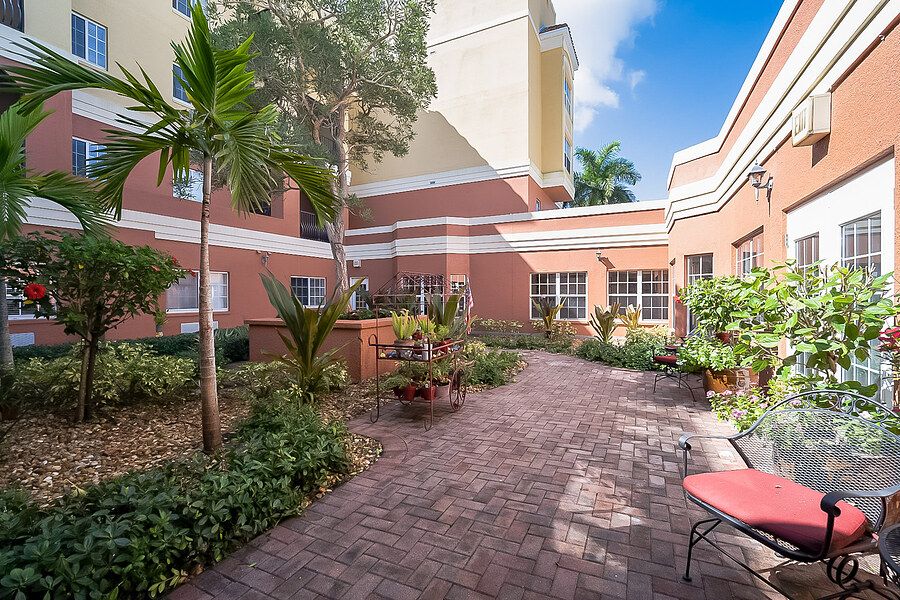A peaceful courtyard with a brick pathway, surrounded by lush plants and a seating area, adjacent to a residential building on a sunny day.