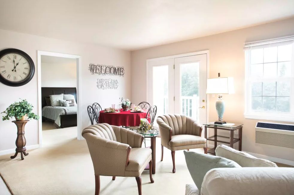 A cozy living room with elegant furnishings, featuring plush chairs, a table set for dining, and a soft lamp lighting the space.