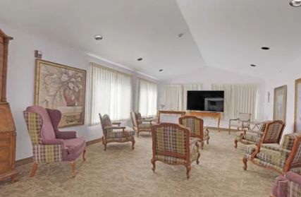 A spacious living room with several chairs arranged around a television and large windows with curtains.