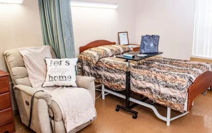 A cozy room with a hospital bed and an overbed table, featuring an armchair with a decorative pillow.