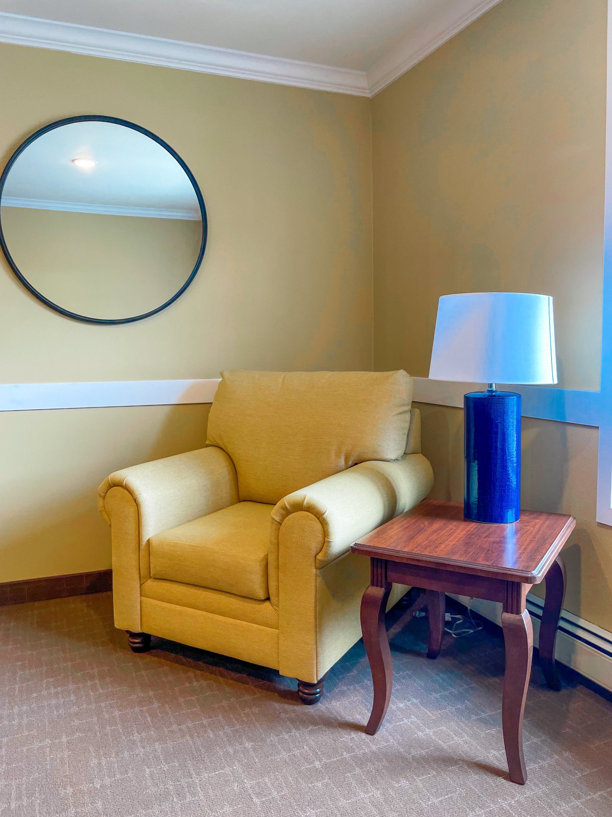 A cozy corner featuring a yellow armchair, round mirror, and a wooden side table with a blue lamp.