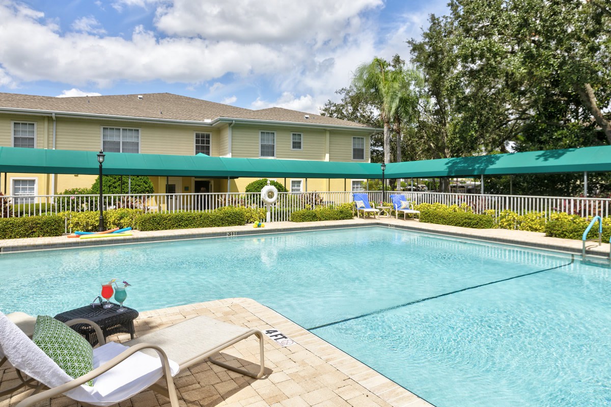 A tranquil residential swimming pool area surrounded by greenery and a two-story building under a clear, sunny sky.
