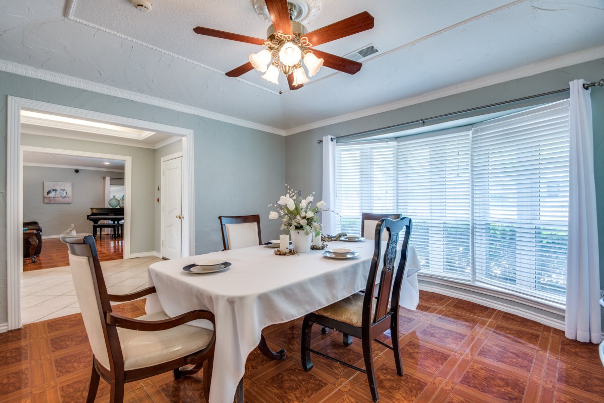 A well-lit dining room featuring a table set for four, with a ceiling fan above and large windows with blinds.