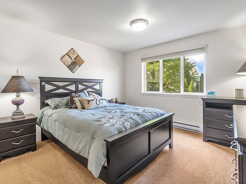 A cozy bedroom featuring a large bed with pillows, a nightstand, and a window with a view of greenery.