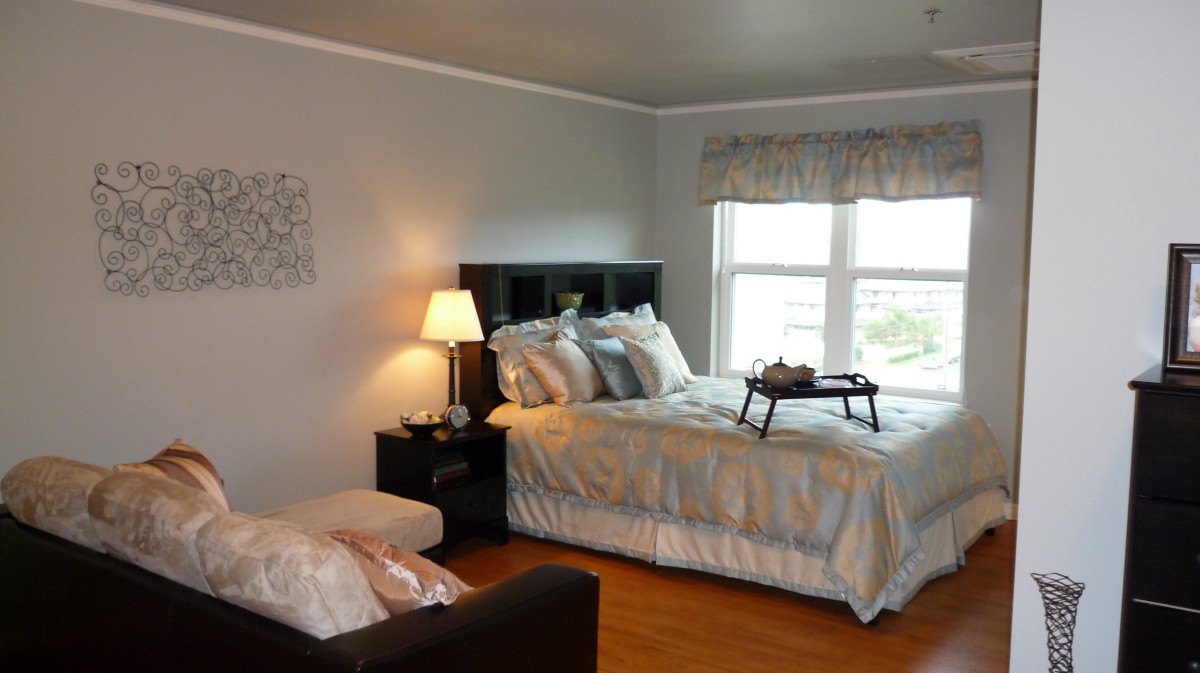 A cozy bedroom with a neatly made bed, bedside lamp, and a window letting in natural light.