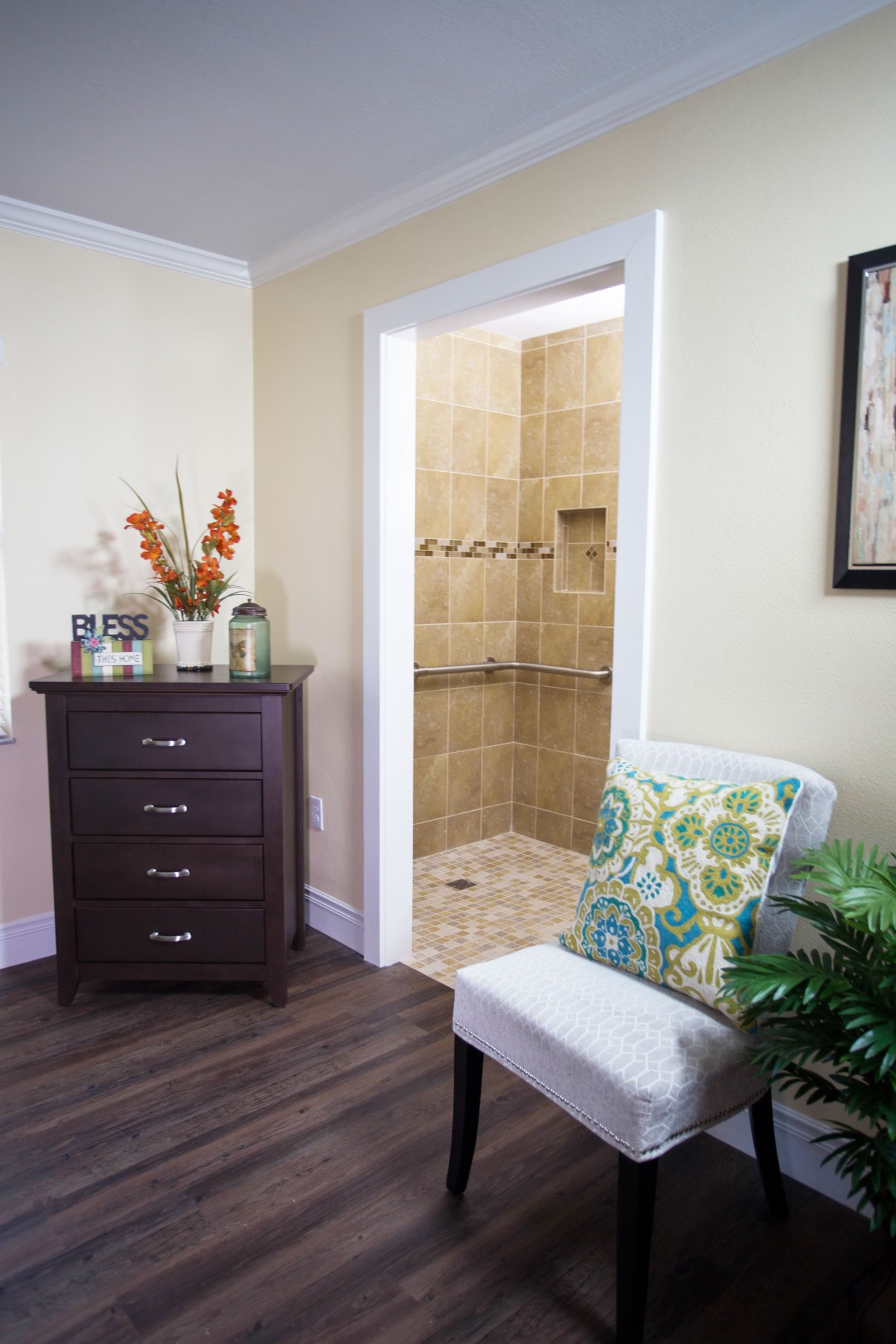 A cozy living space featuring a wooden drawer and comfortable chair with a floral pillow, leading into a tiled bathroom.
