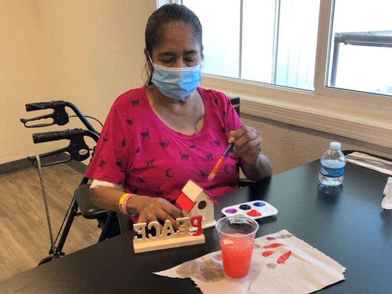 A person wearing a mask is painting a birdhouse at a table.