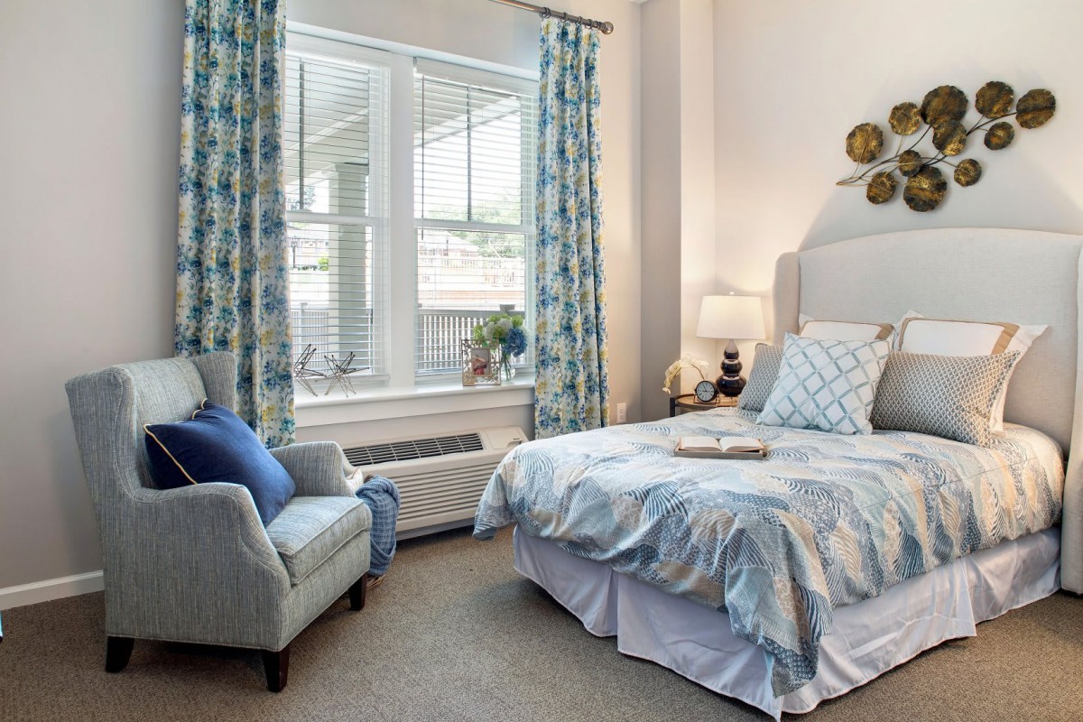 A cozy and well-decorated bedroom featuring a bed with decorative pillows, an armchair by the window, and elegant curtains.