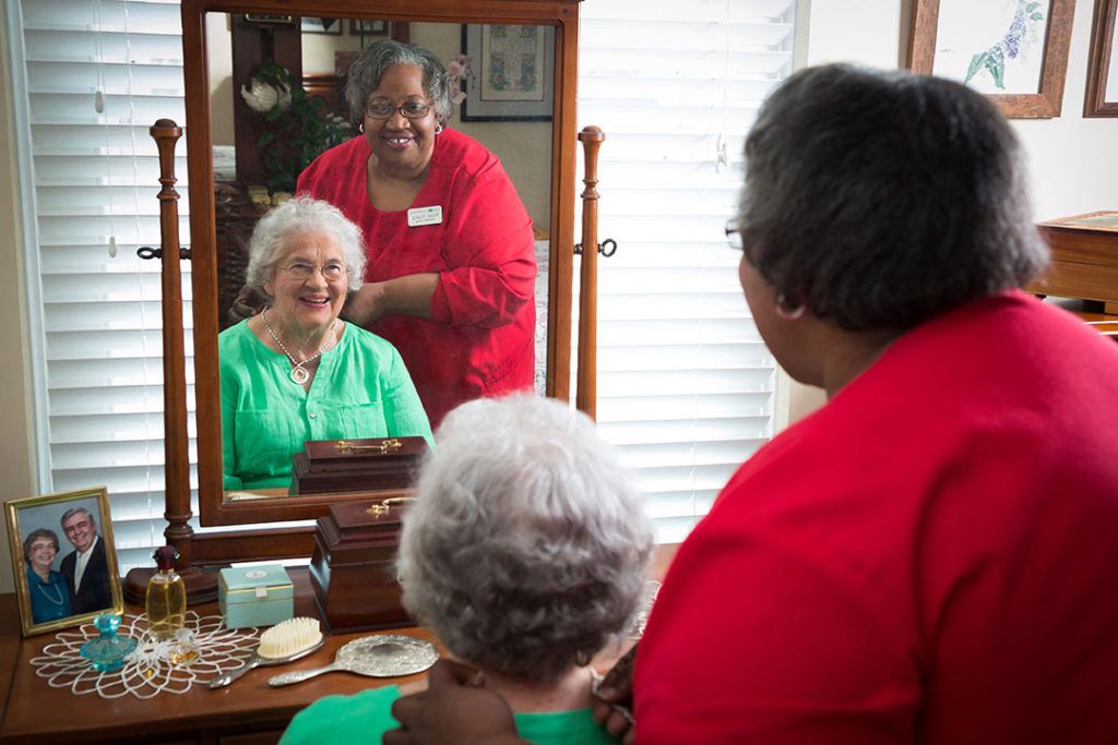 A caregiver assists an elderly woman in front of a mirror, creating a moment of warmth and connection.