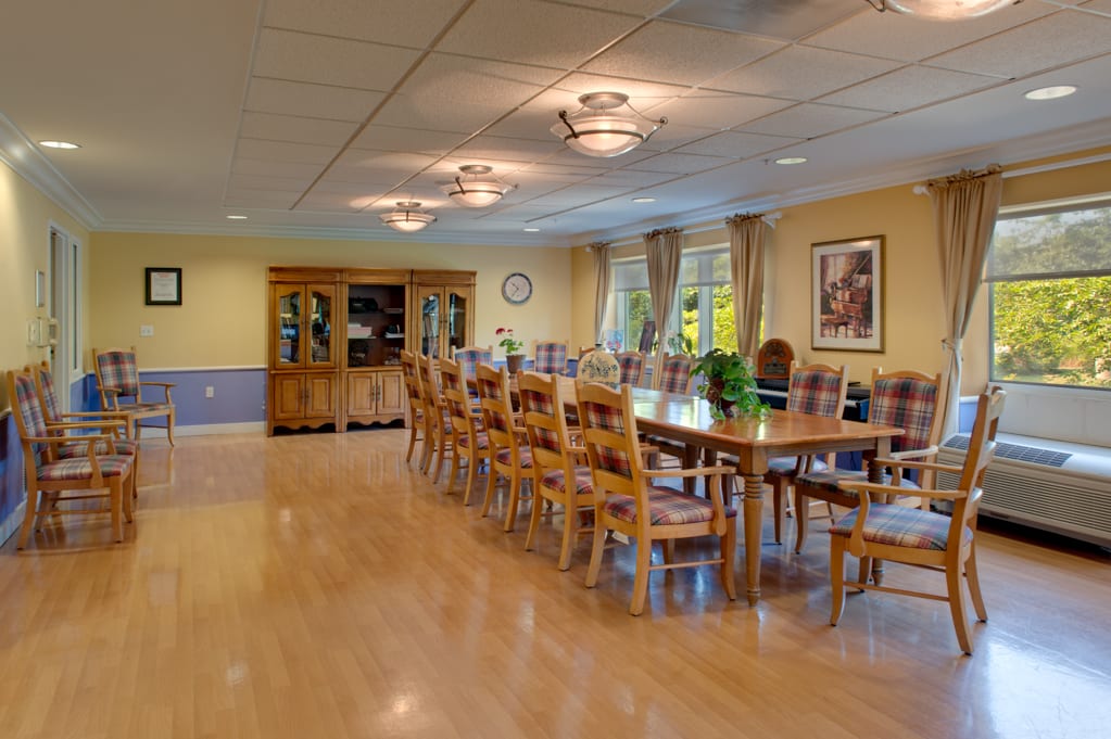 A spacious dining room with a long wooden table and numerous chairs, adorned with decorative lighting and large windows with curtains.