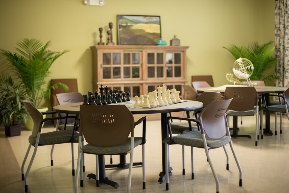 A cozy game room featuring a chessboard setup on a table, surrounded by chairs, with a cabinet and trophies in the background.