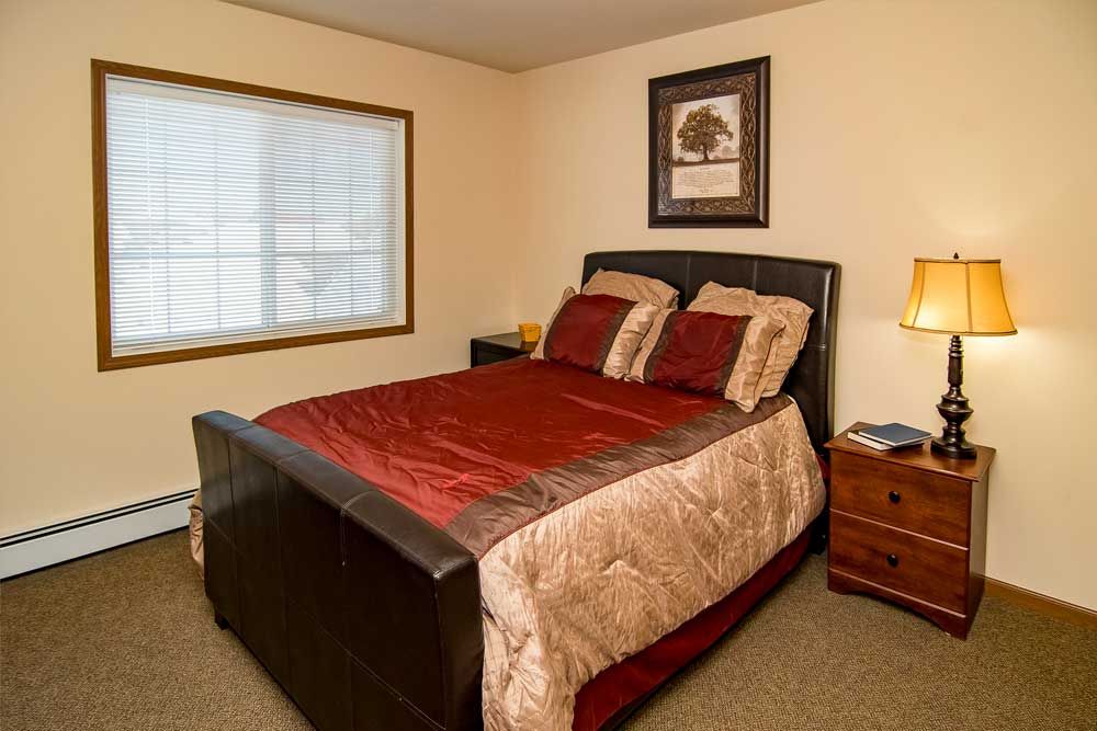 A cozy bedroom featuring a neatly made bed with red and beige bedding, a window, and a nightstand with a lamp.