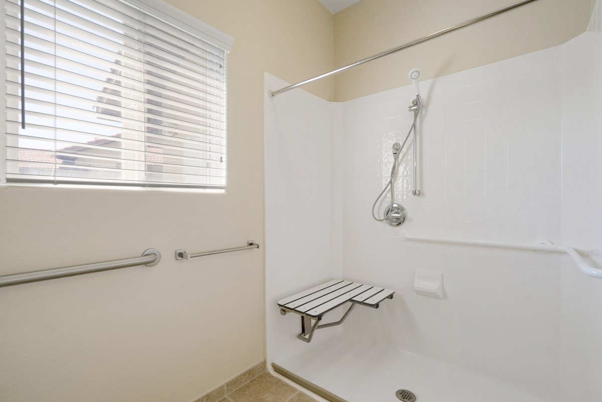 An accessible bathroom featuring a shower with grab bars, a foldable bench, and a handheld showerhead next to a window.