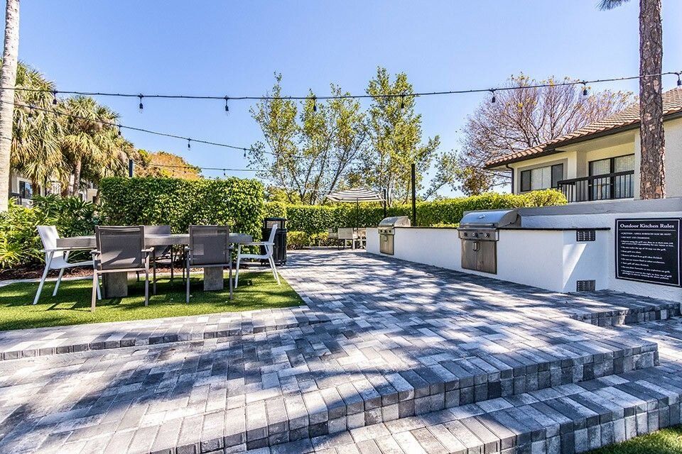 A spacious backyard patio with a dining area and grill, adorned with string lights on a sunny day.
