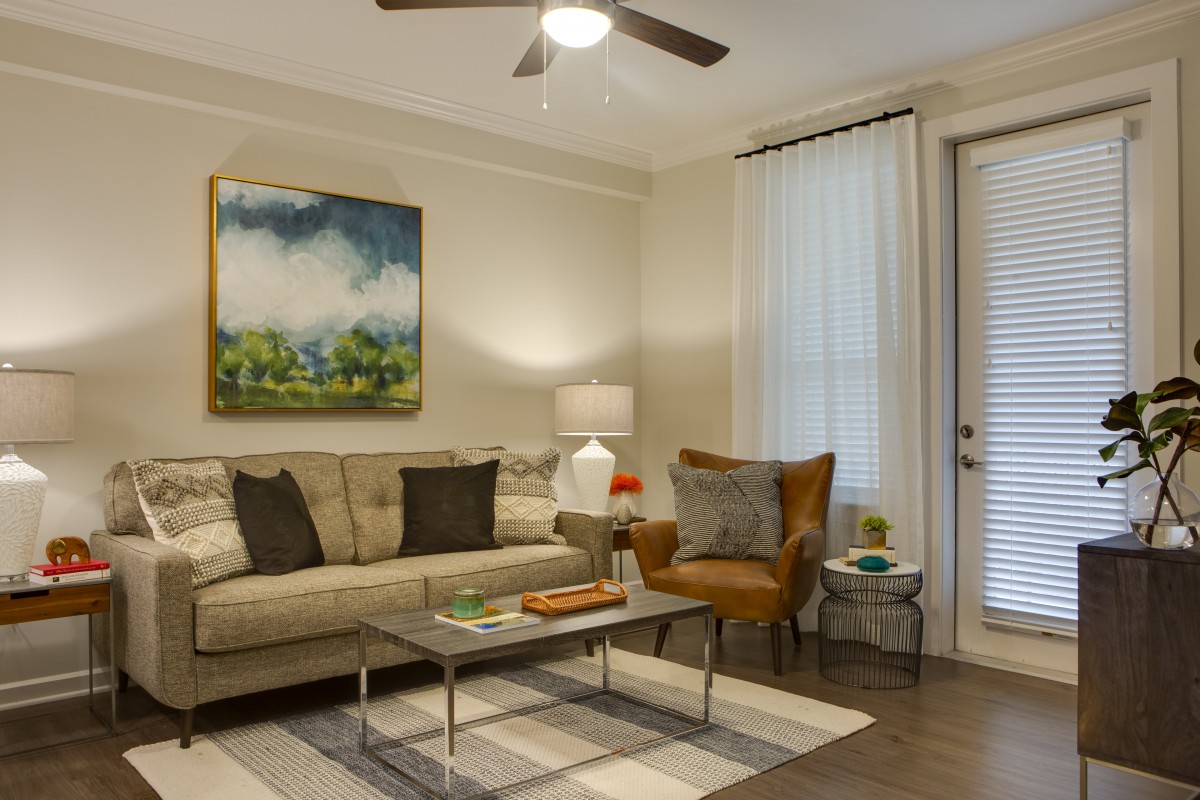 A cozy living room with a sofa, armchair, and coffee table beneath a ceiling fan, decorated with a colorful painting.
