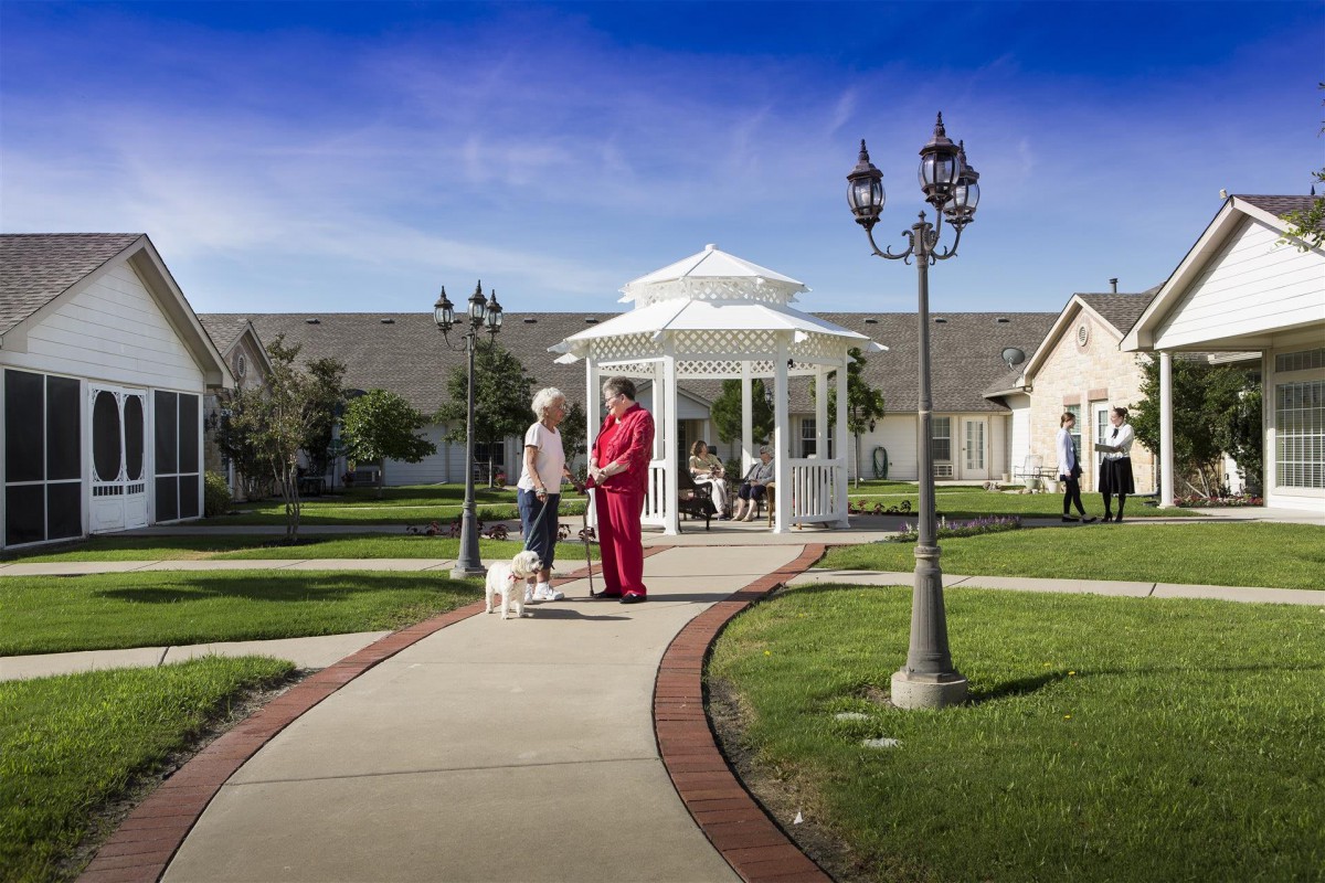 Two people walking with a dog near a gazebo on a sunny day in a nursing home garden.