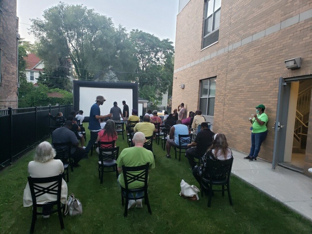 An outdoor event with an audience seated in front of a screen near a brick building.